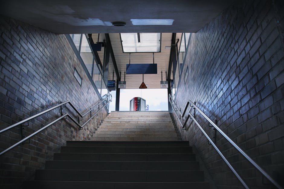 A set of yellow and black striped metal stairs inside a station or industrial building, with yellow handrails on both sides and a sign on one of the steps reading 'PLEASE KEEP LEFT.' The stairs lead u